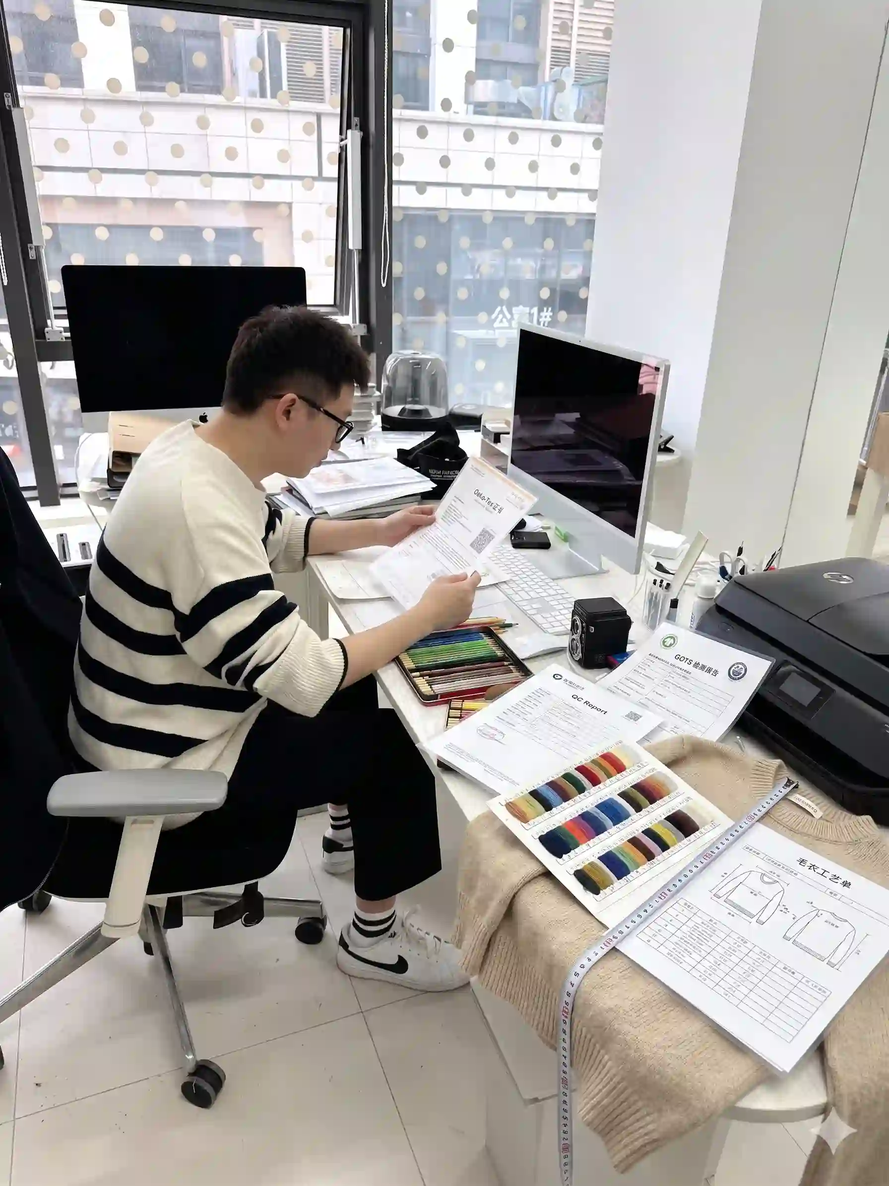 Real office shot of merchandiser reviewing OEM sweater tech pack, yarn color card, size spec sheet and GOTS certification, custom knitwear design & development workflow photo