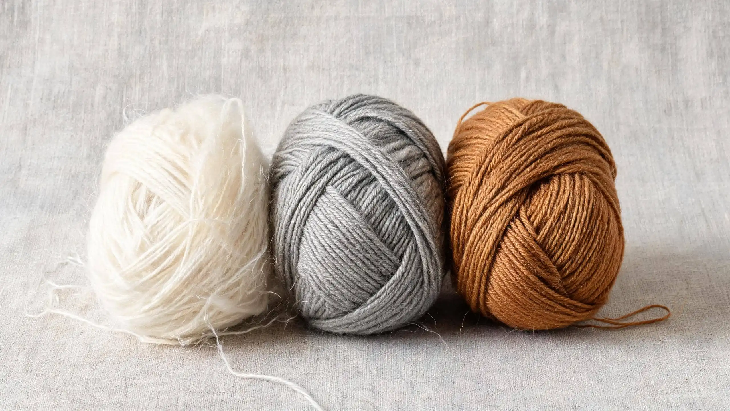 Three yarn skeins side by side comparison: white cashmere, gray merino wool, and brown alpaca on linen background