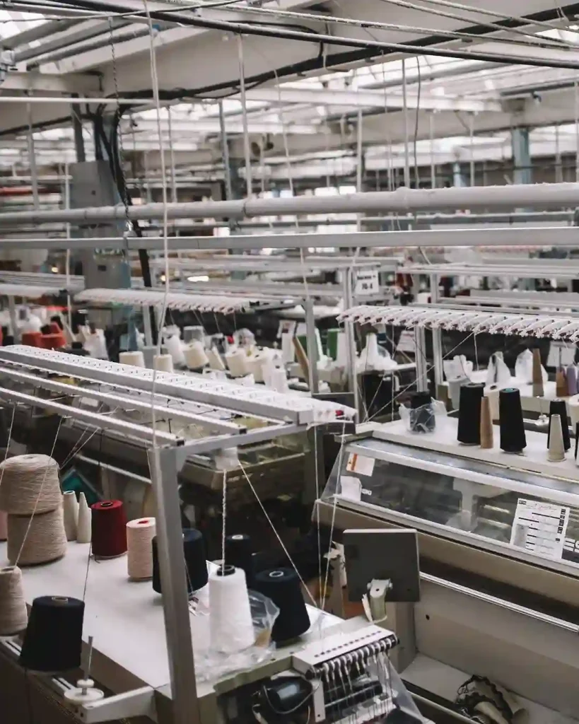 Modern sweater OEM factory floor with industrial knitting machines and colorful yarn cones, showcasing advanced knitwear production technology.