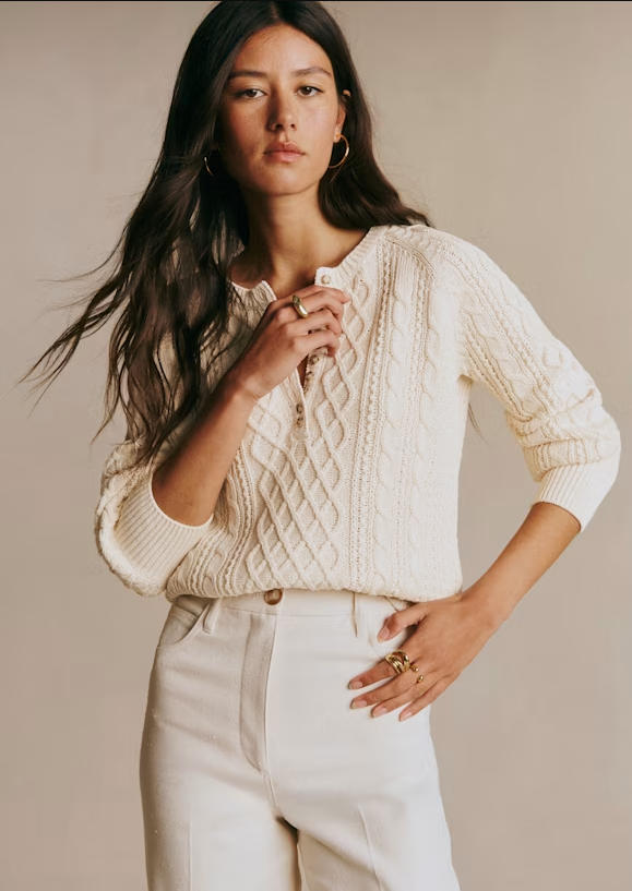 Studio full-body fashion shot of cream twisted organic cotton henley cable knit top, diamond cable stitch raglan sleeve organic knit pullover, minimalist neutral tone women daily knitwear matched with white trousers